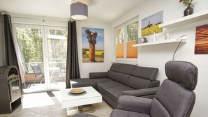 Een moderne woonkamer met een grijze bank, een witte salontafel en een grijze poef. Links bevindt zich een open haard, een raam met zonwering en een schilderij aan de muur.
