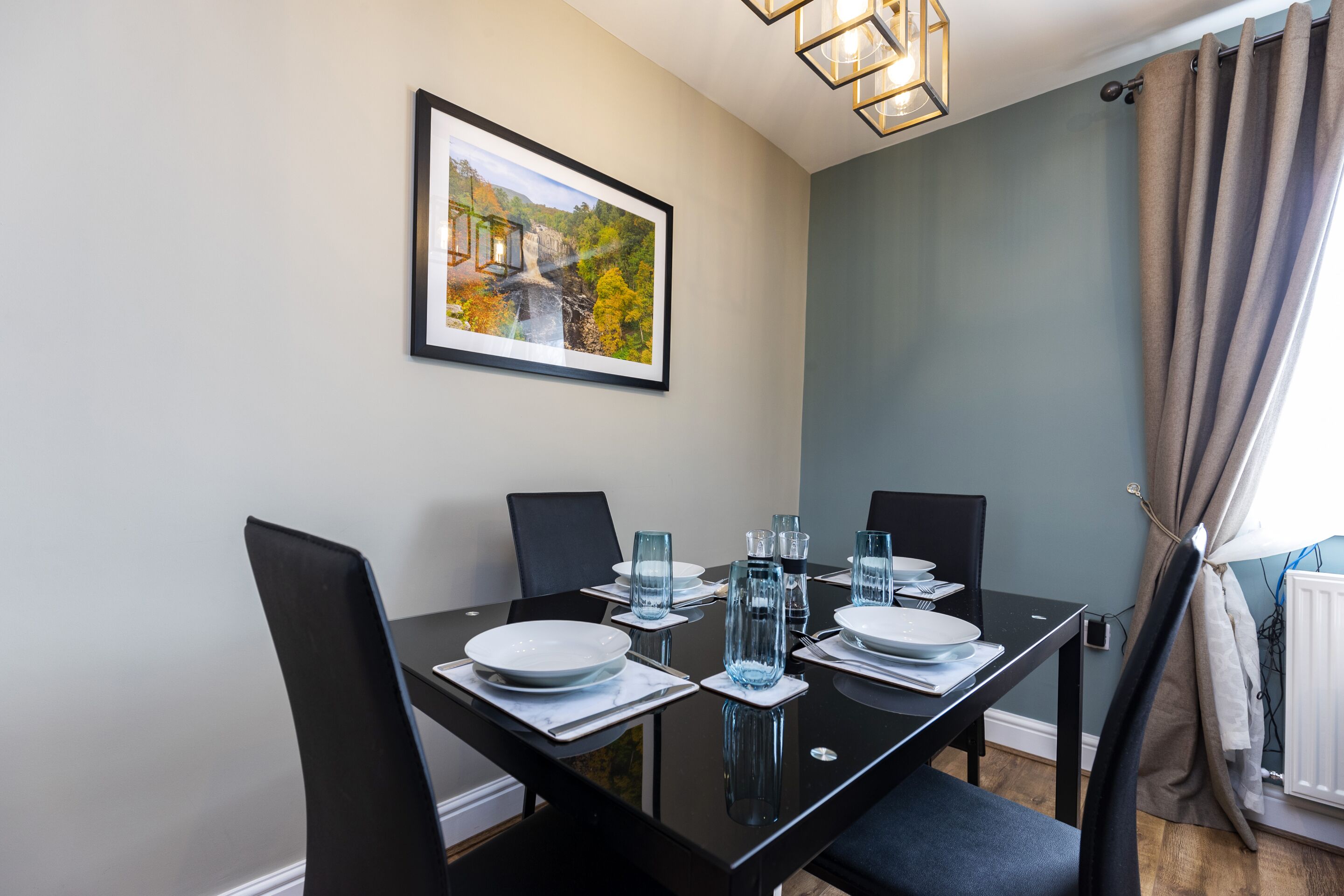 A dining area with a black table set for four, including plates, glasses, and cutlery. There is a framed picture on the wall above the table.