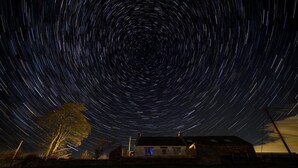 Fotografía de estelas de estrellas con una casa y un árbol en primer plano.