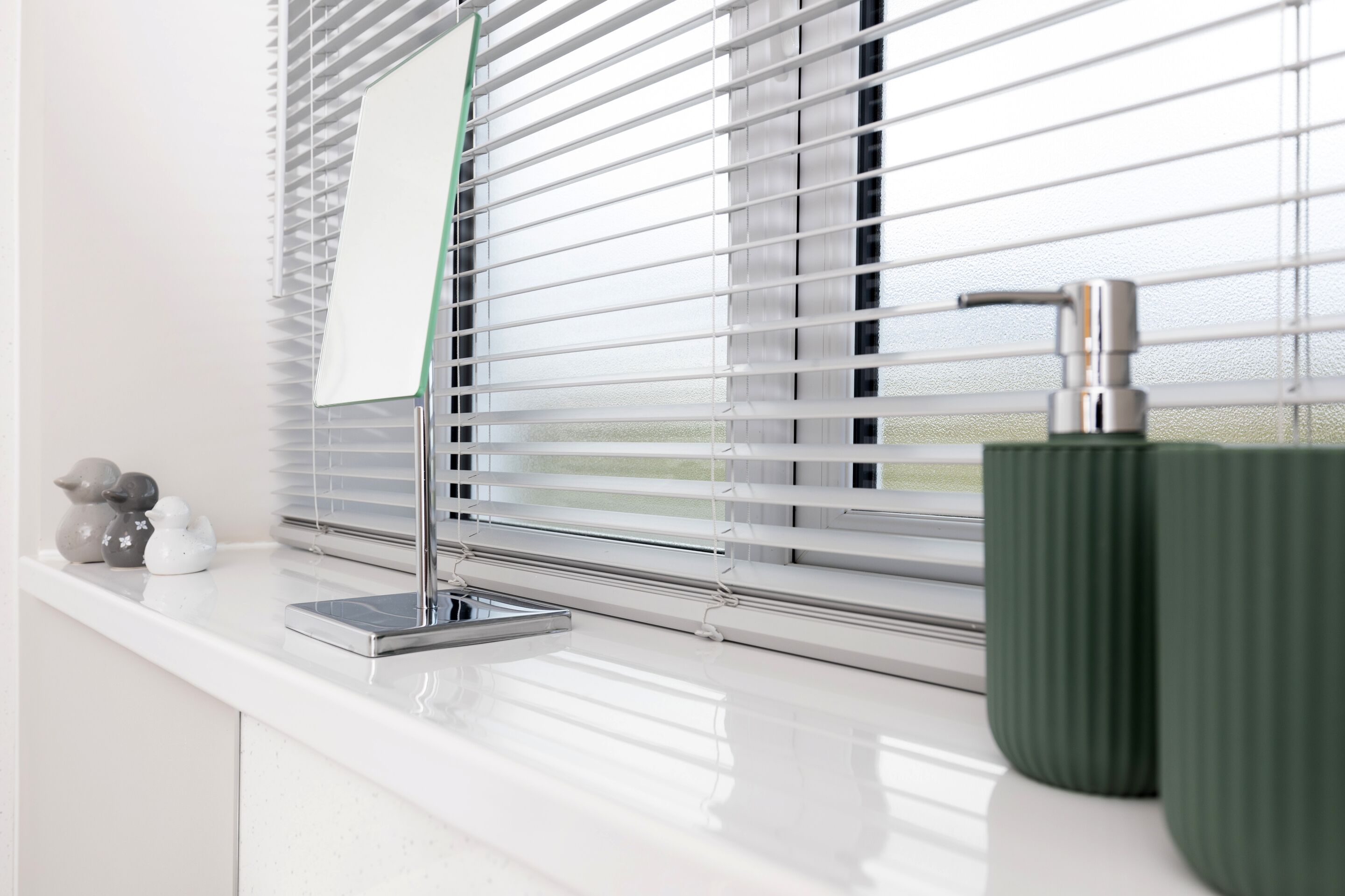 A bathroom with a white countertop, a green vase, and a window with blinds.