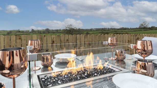Un espacio de comedor al aire libre con una mesa de vidrio, copas de vino de cobre y una chimenea.