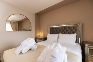 Habitación de hotel con una cama grande, un espejo redondo, una mesita de noche con lámpara y dos toallas con la palabra 'slippers'.