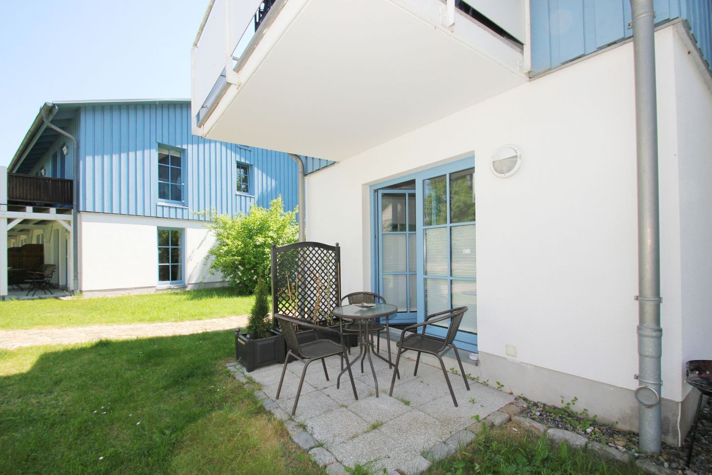 A patio with a table and chairs, a small garden, and a building with blue windows and a white exterior.