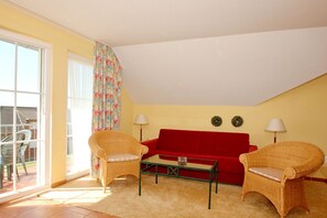 A cozy living room with a red sofa, two wicker chairs, a glass coffee table, and a large window with a curtain. - Plenty of nature, an idyllic setting, a quiet location - Ferienpark Neddesitz FeWo 48 - (Sagard)