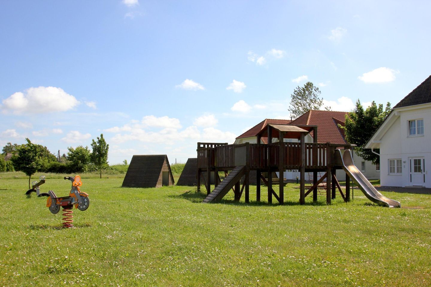 Ein Spielplatz mit Rutsche und Klettergerüsten, umgeben von einer grünen Wiese und einem Gebäude mit rotem Dach.