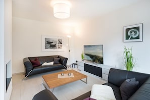 En moderne stue med en sort lædersofa, et træ sofabord og en sort lædersofa. Der er et fladskærms-tv monteret på væggen, en gulvlampe og et indrammet billede af en reveplante.