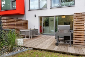 En moderne udendørs terrasse med træterrasse, skydedør i glas, grill og spisebord med stole.