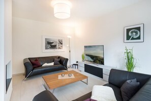 Un salon moderne avec un canapé en cuir noir, une table basse en bois et un divan en cuir noir. Un téléviseur à écran plat est fixé au mur, ainsi qu’une lampe sur pied et un tableau encadré représentant un pissenlit.
