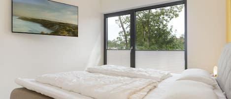 A bedroom with a large window, a bed with white bedding, and a painting on the wall.