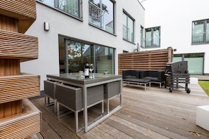 Een terras buiten met een tafel met glazen blad, rieten meubels en een barbecue.