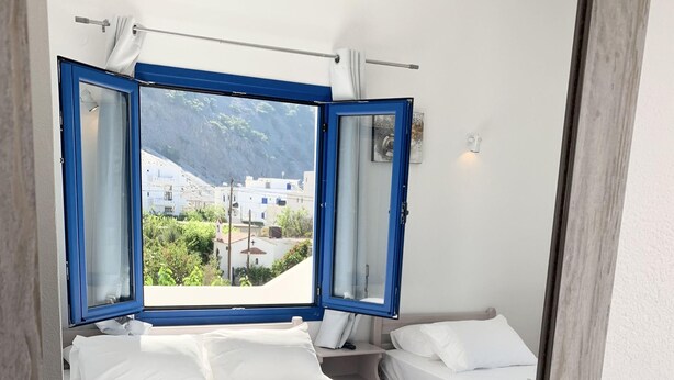 A hotel room with a bed, a bedside table, a mirror, and a view of the outside. - Giannis Rooms (Chora Sfakion)