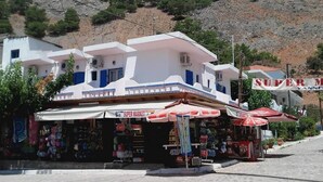 A white building with a blue door and windows, a sign reading "Super Market", and a red umbrella. - Giannis Rooms (Chora Sfakion)