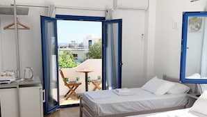 A hotel room with two beds, a small kitchenette, a dining area with a table and chairs, and a balcony with a view of buildings and greenery. - Giannis Rooms (Chora Sfakion)
