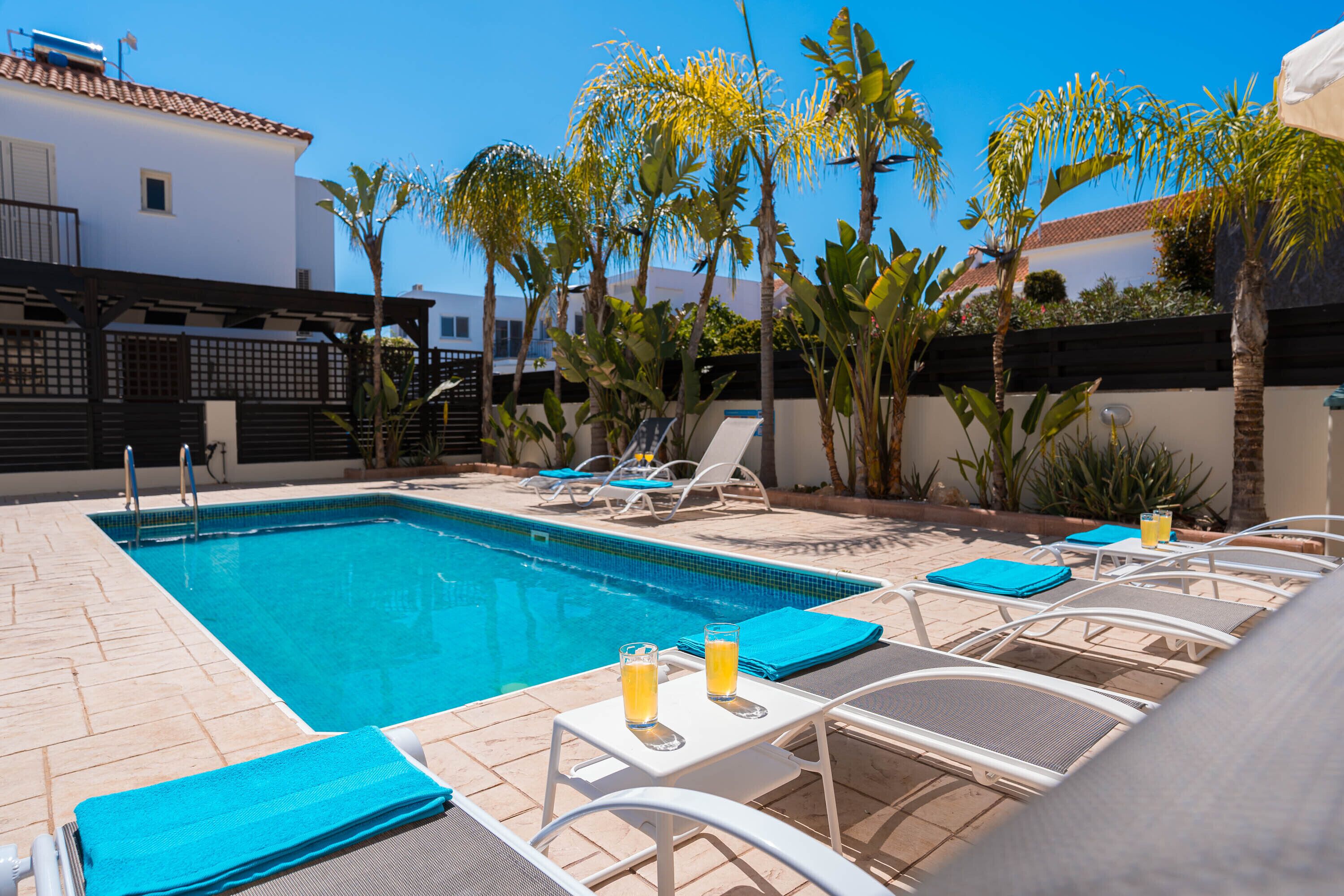 A swimming pool with lounge chairs and umbrellas, surrounded by palm trees and a white building.