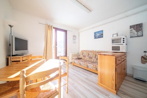A living room with a wooden dining table, chairs, a sofa, a television, and a microwave. - L'Ourson - Studio avec balcon plein sud (Les Angles)