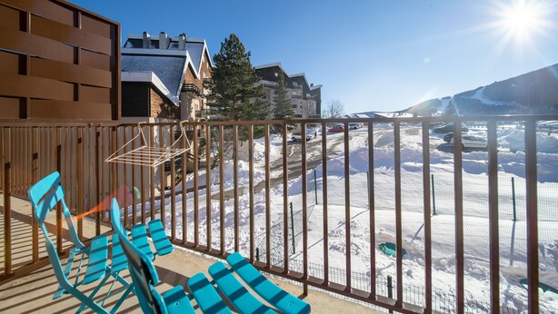 Un balcon avec des chaises bleues, une balustrade en bois et une vue sur un paysage enneigé, avec des bâtiments en arrière-plan.