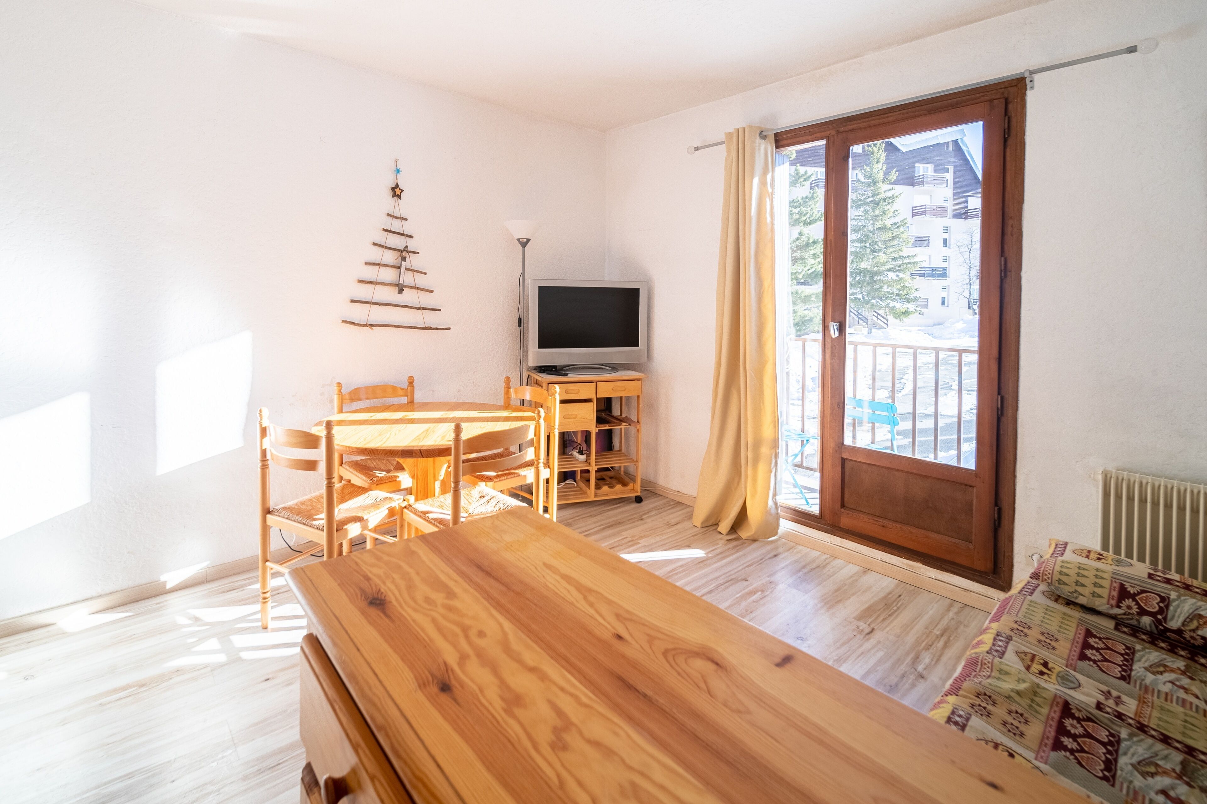 Une pièce avec une table en bois, des chaises, un téléviseur posé sur un meuble, une fenêtre avec des rideaux et une vue sur les bâtiments à l’extérieur.