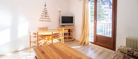 Une pièce avec une table en bois, des chaises, un téléviseur posé sur un meuble, une fenêtre avec des rideaux et une vue sur les bâtiments à l’extérieur.
