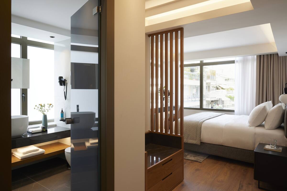 Quarto de hotel moderno, com uma janela grande, uma cama, uma mesa de cabeceira e um espaço de banheiro com pia e espelho.