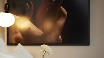 A framed artwork on a wall, a bedside lamp, a white flower, and a bed with white bedding.