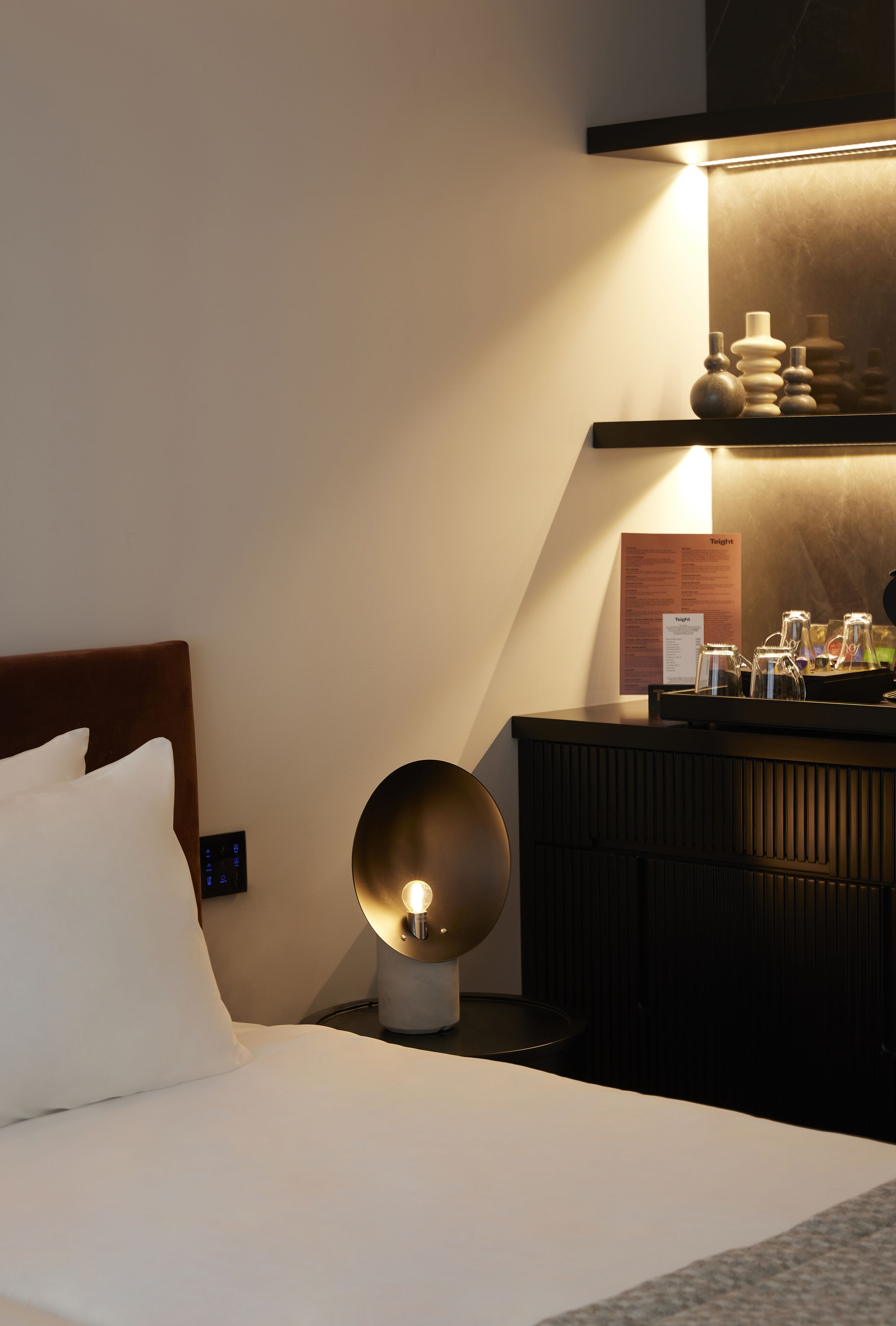 A hotel room with a bed, a bedside table with a lamp, a nightstand, and a wall-mounted shelf with decorative items.