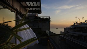 A balcony with a view of the sea at sunset, a potted plant, and a white curtain.