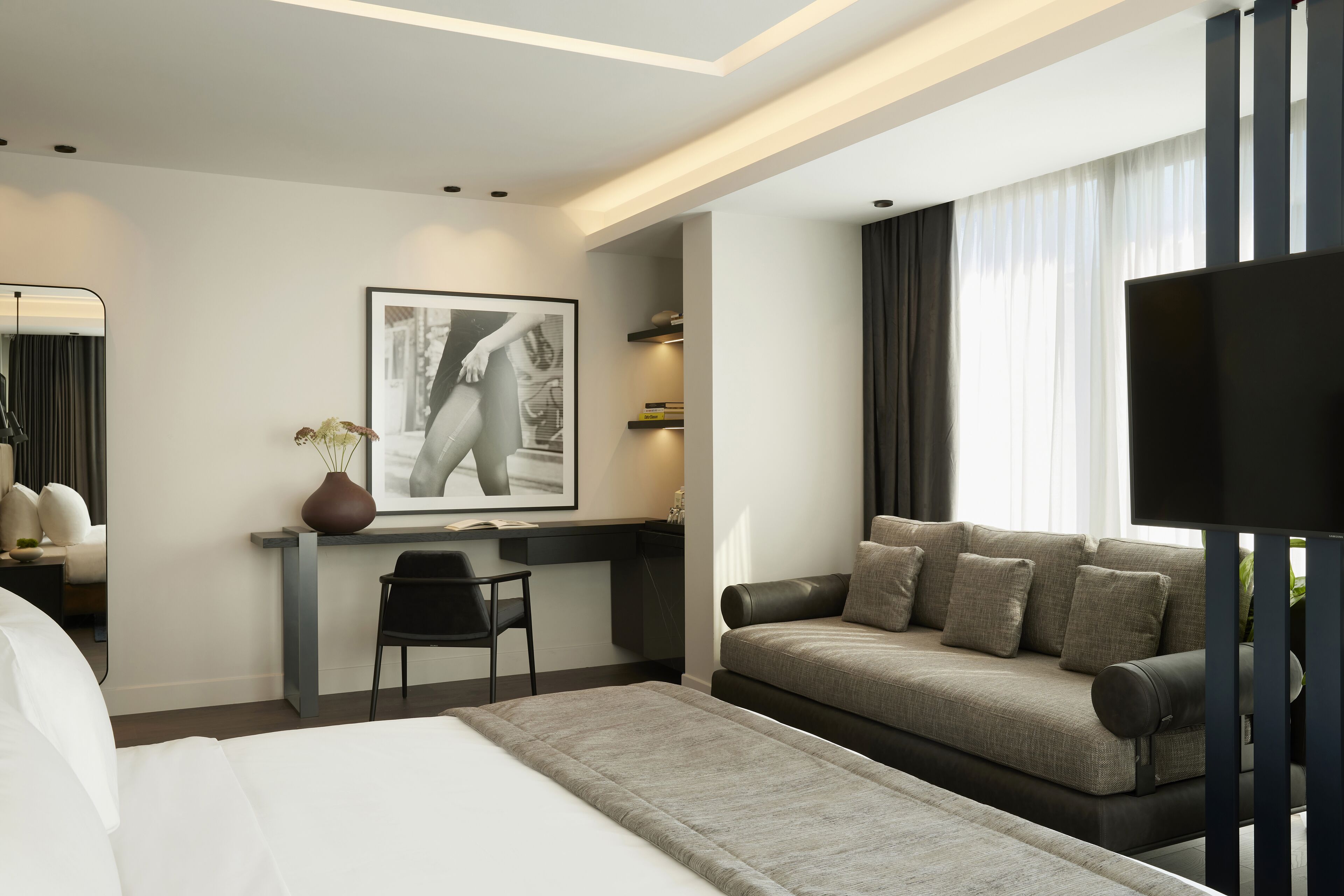 A modern hotel room with a bed, a desk, a chair, a sofa, and a TV.