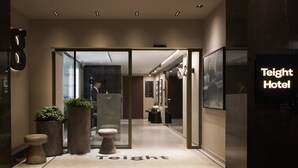 A hotel entrance with a modern design, featuring a reception desk, potted plants, and a clear view into the lobby.