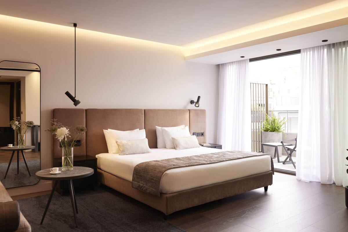 Quarto de hotel moderno com uma cama grande, mesas de cabeceira, um espelho e uma varanda com plantas.