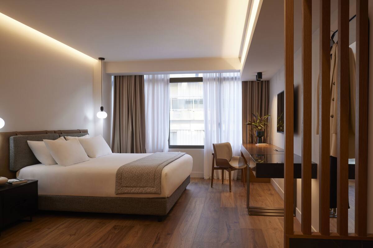 Quarto de hotel com uma cama grande, uma escrivaninha, uma cadeira, televisão e janela com cortinas.