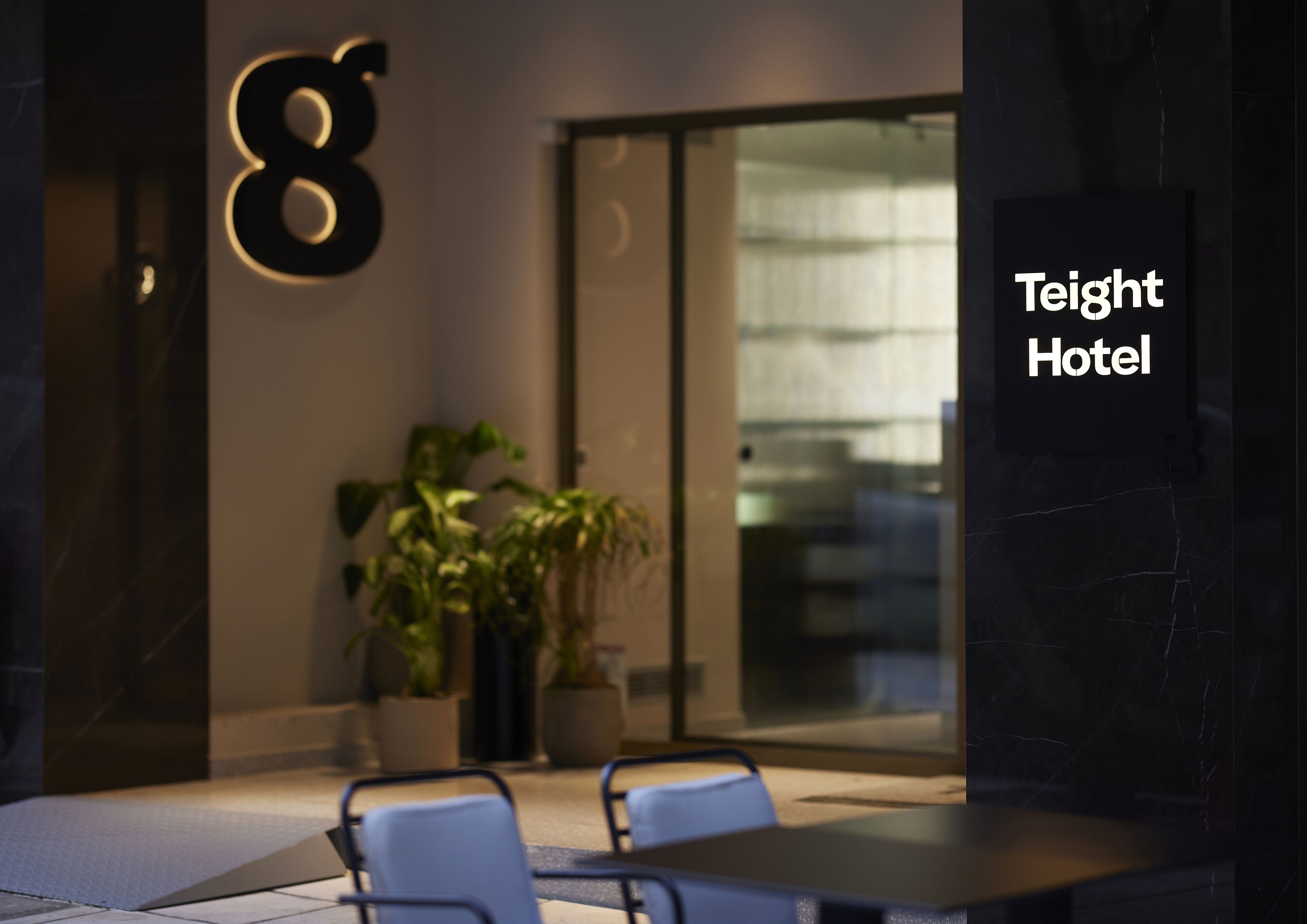 Entrada del Teight Hotel con un diseño moderno, que destaca un gran número '8' iluminado en la pared, plantas en macetas y una zona de estar con sillas y una mesa.