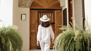 Una persona caminando por un pasillo con pared de ladrillo rústico, puertas de madera y helechos en macetas.