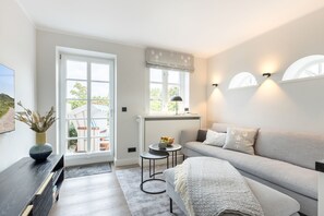 Ein modernes Wohnzimmer mit einem grauen Sofa, einem kleinen runden Tisch mit Obstschale, einem dunklen Holznachttisch und einer Vase mit Trockenpflanzen.