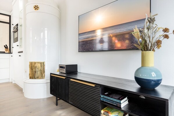 A modern living room with a black entertainment unit, a large painting of a sunset over the ocean, and a vase with dried flowers. - Friesenhof App. 06 - Friesenhof (Sylt)