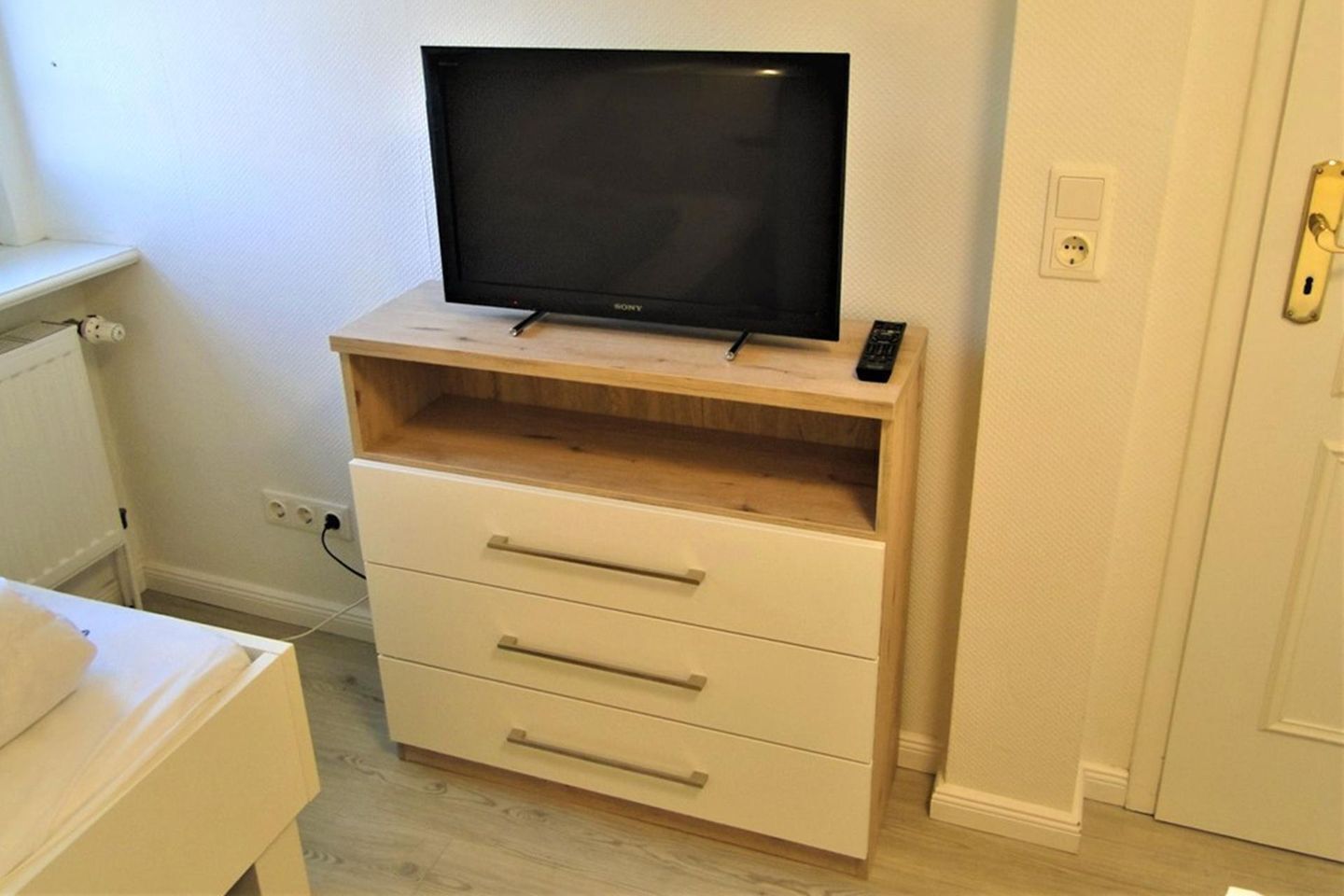 Ein Zimmer mit Fernseher auf einer Holzkassette, einer weißen Kommode mit Schubladen, einem weißen Heizkörper und einer Tür mit Schloss.