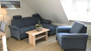 En moderne loftstue med en mørkeblå sofa, en matchende lænestol og et træbord. Et indrammet landskabsmaleri hænger på væggen, og et vindue giver naturligt lys.