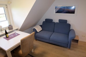 A room with a blue sofa, a small table with a bottle and glass, and a framed picture on the wall. - Hüs Thora Jaiken "App.Birthe" TJ/02 - Thora Jaiken (Sylt)