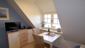 Ein kleiner, gemütlicher Raum mit schrägen Wänden, einem Schreibtisch mit Stuhl, einem Fernseher, einem Fenster mit Aussicht und einer kleinen Pflanze.