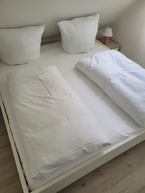 A neatly made bed with white bedding and pillows in a room with wooden flooring and a nightstand with a lamp. - Hüs Thora Jaiken "App.Birthe" TJ/02 - Thora Jaiken (Sylt)