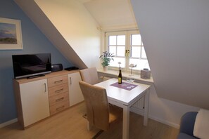 Ein kleiner, gemütlicher Raum mit schrägen Wänden, einem Schreibtisch mit Stuhl, einem Fernseher, einem Fenster mit Aussicht und einer kleinen Pflanze.