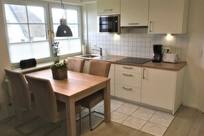 A modern kitchen with a dining area, featuring a wooden table, chairs, a black pendant light, a microwave, a toaster, and a coffee maker. - Hüs Thora Jaiken "App. Eike" TJ/03 - Thora Jaiken (Sylt)