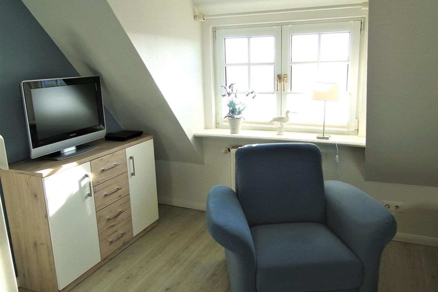 A room with a television on a wooden cabinet, a blue chair, and a window with a plant.