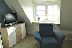 A room with a television on a wooden cabinet, a blue chair, and a window with a plant. - Hüs Thora Jaiken "App. Eike" TJ/03 - Thora Jaiken (Sylt)