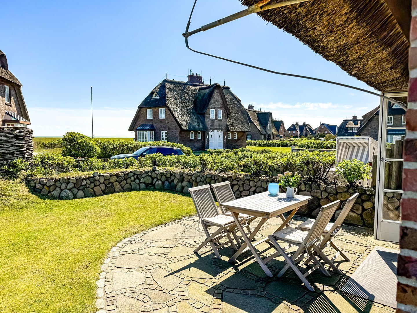 A stone patio with outdoor furniture, a thatched-roof house, and a stone wall in the background.
