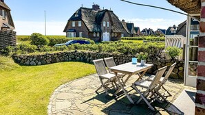 En stenbelagt terrasse med udendørs møbler, et stråtækt hus og en stenvæg i baggrunden.