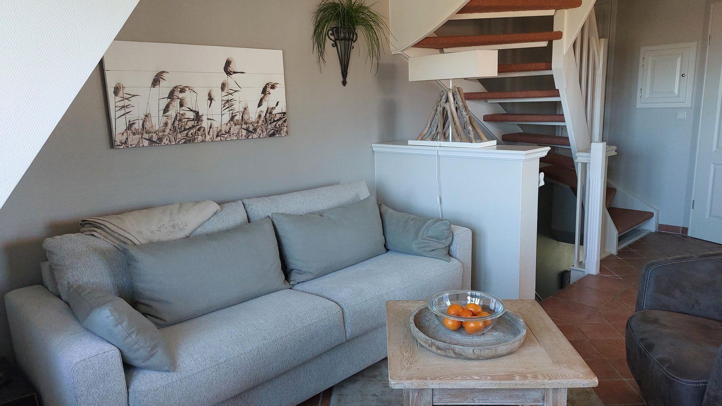Un salon avec un canapé gris, une table basse en bois sur laquelle se trouve un saladier rempli de fruits, un meuble blanc et un escalier avec des marches rouges.