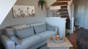 Un salon avec un canapé gris, une table basse en bois sur laquelle se trouve un saladier rempli de fruits, un meuble blanc et un escalier avec des marches rouges.