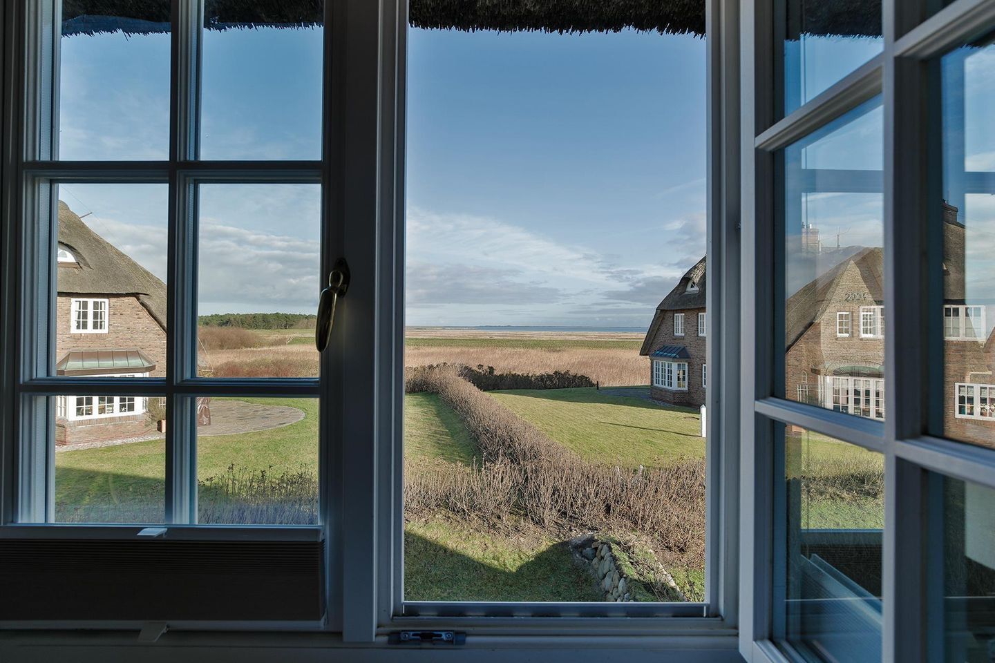 A view through a window with a clear sky, a grassy field, and two brick houses with thatched roofs.