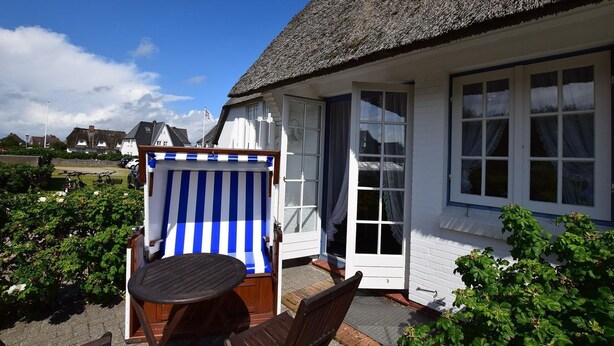 A white building with a thatched roof, a covered outdoor seating area with a striped awning, and a view of the surrounding landscape. - Wattstübchen SO/16a - Söl'ring Hüs (Sylt)
