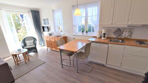 A modern kitchen with white cabinets, a wooden dining table, and a window with curtains. - Wattstübchen SO/16a - Söl'ring Hüs (Sylt)